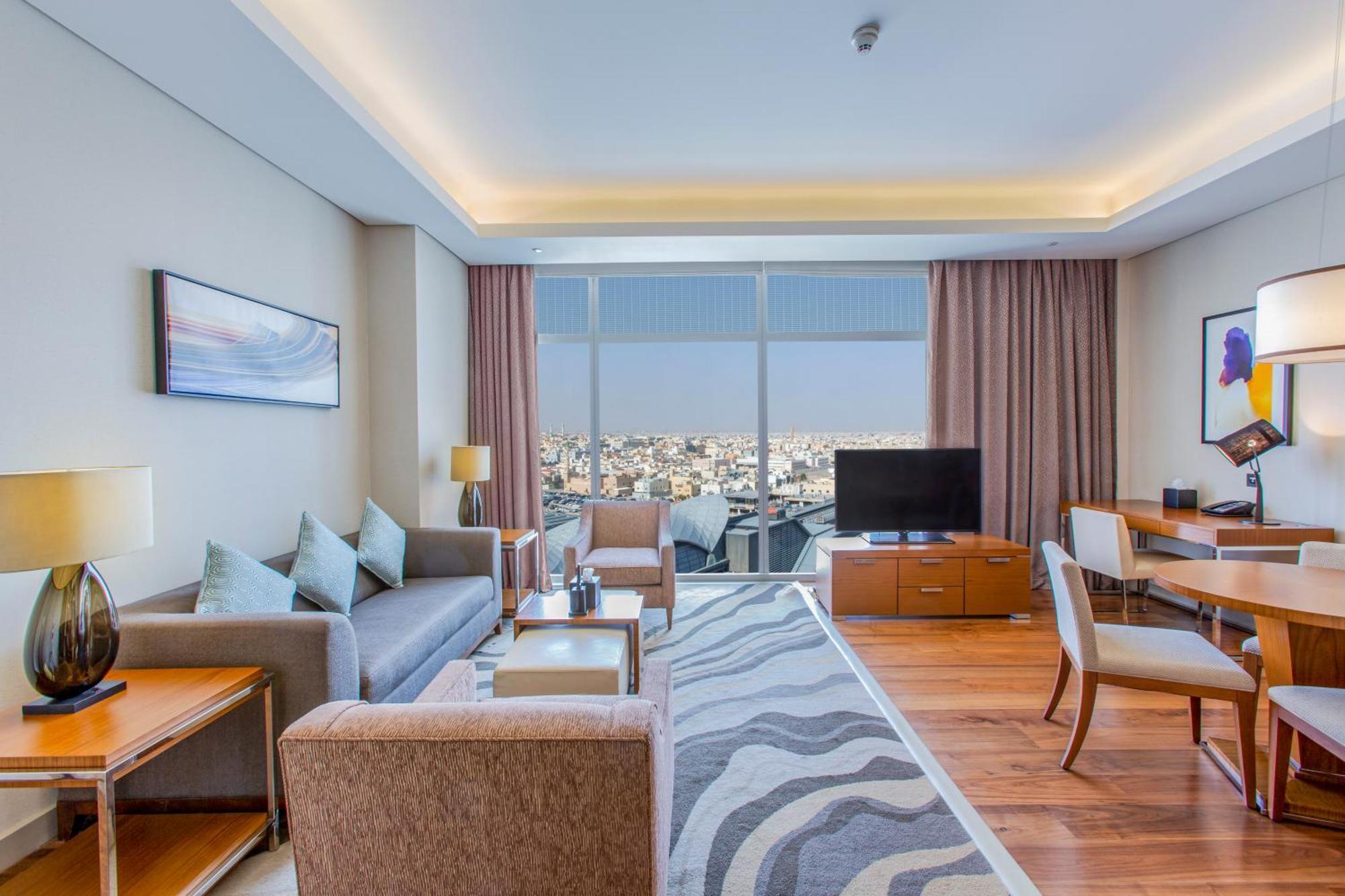 Hilton Riyadh Hotel & Residences Eksteriør billede The photo shows a modern hotel room or suite. The space features a stylish living area with a sofa and armchairs arranged around a coffee table. There are lamps providing ambient lighting, and a large window offers a view of the cityscape outside. A 
