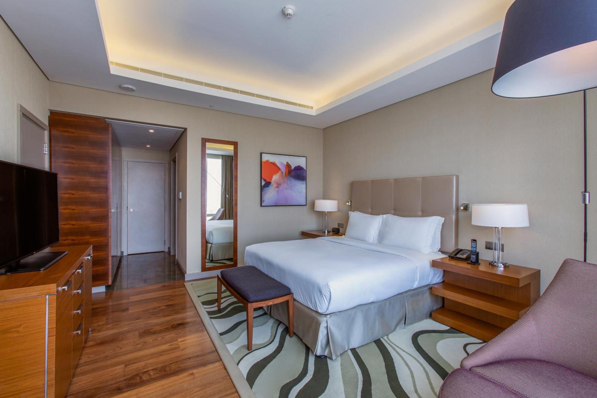 Hilton Riyadh Hotel & Residences Eksteriør billede The photo shows a modern hotel room with a comfortable and stylish design. It features a large bed with white linens and a headboard, flanked by two bedside lamps. There is a small bench at the foot of the bed and a cozy chair in the corner. A flat-s