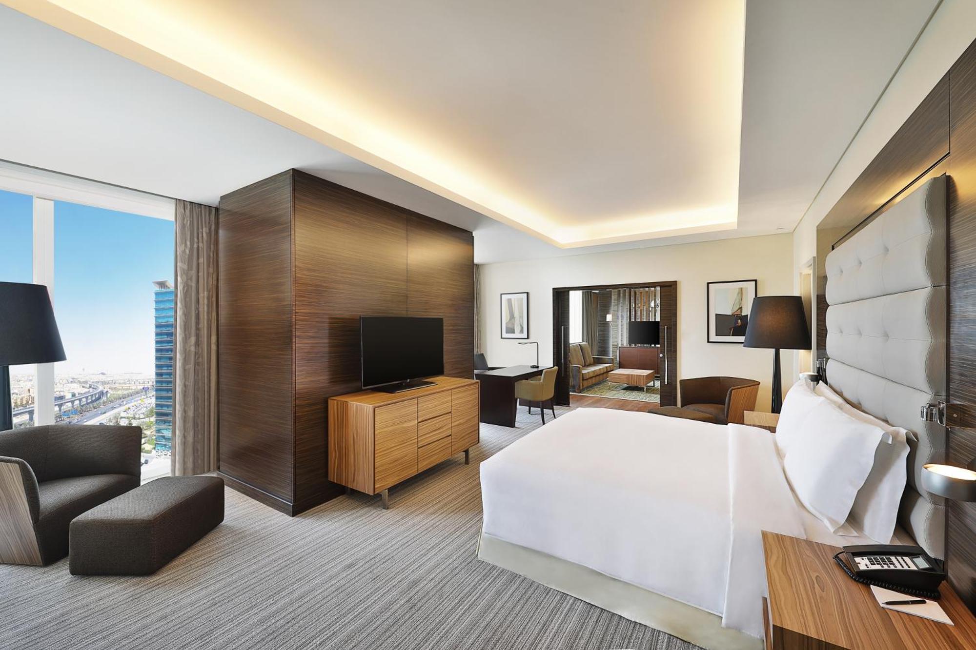 Hilton Riyadh Hotel & Residences Eksteriør billede The photo shows a modern hotel room. It features a neatly made bed with white linens, a wooden nightstand beside it, and a cozy armchair. The room has a large window that provides a view of the outside, likely showcasing a cityscape. A flat-screen TV