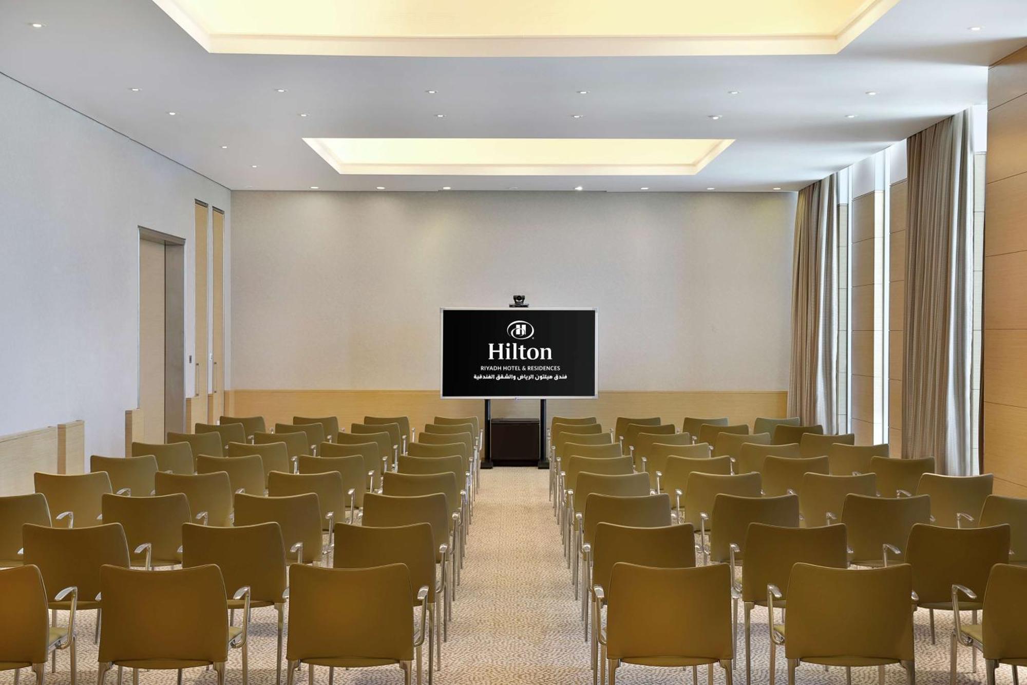 Hilton Riyadh Hotel & Residences Eksteriør billede The photo shows a conference room or meeting space. It features rows of green chairs arranged facing a large screen that displays the Hilton logo. The room has a modern design with light-colored walls and a bright ceiling. Natural light appears to co