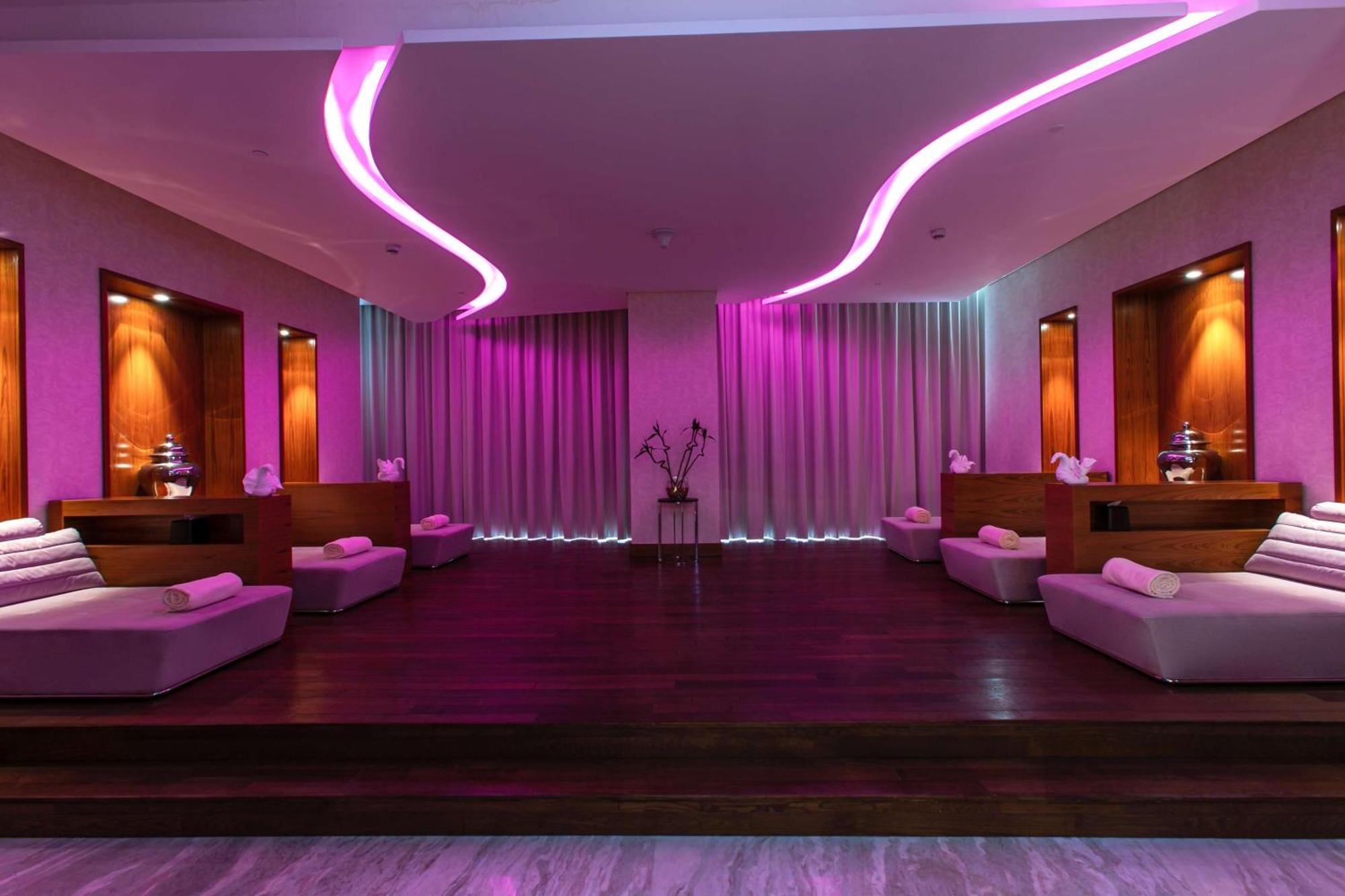 Hilton Riyadh Hotel & Residences Eksteriør billede The photo depicts a relaxing interior space, likely a spa or wellness area. It features a wooden floor with a raised platform accented by soft, low couches on either side. The walls are adorned with modern lighting, which emits a calming pink hue. Th