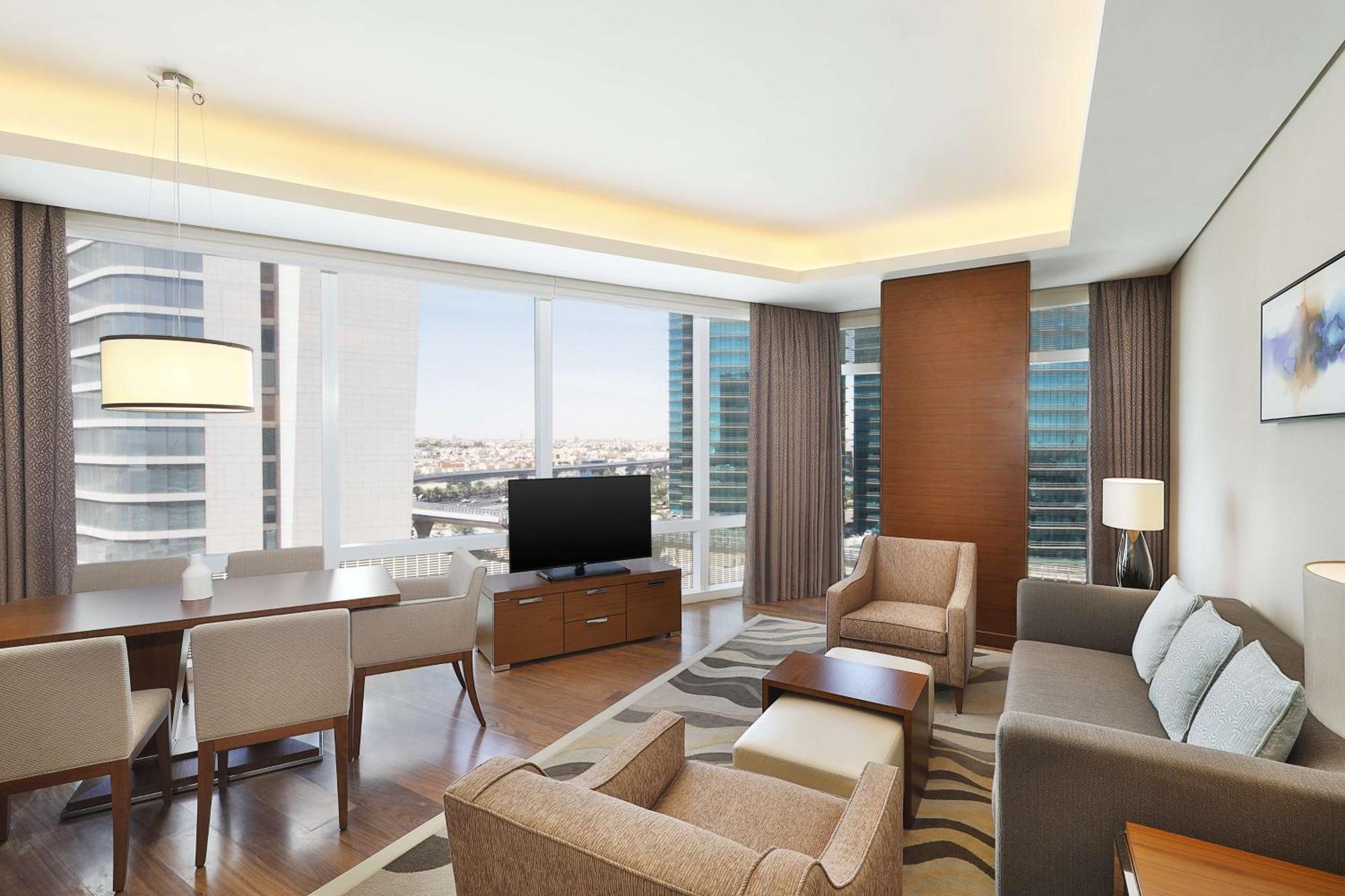 Hilton Riyadh Hotel & Residences Eksteriør billede The image shows a modern, stylish living area in an apartment or hotel suite. It features large windows that provide a view of the cityscape, allowing natural light to flood the space. The furnishings include a comfortable sofa, armchairs, and a coff
