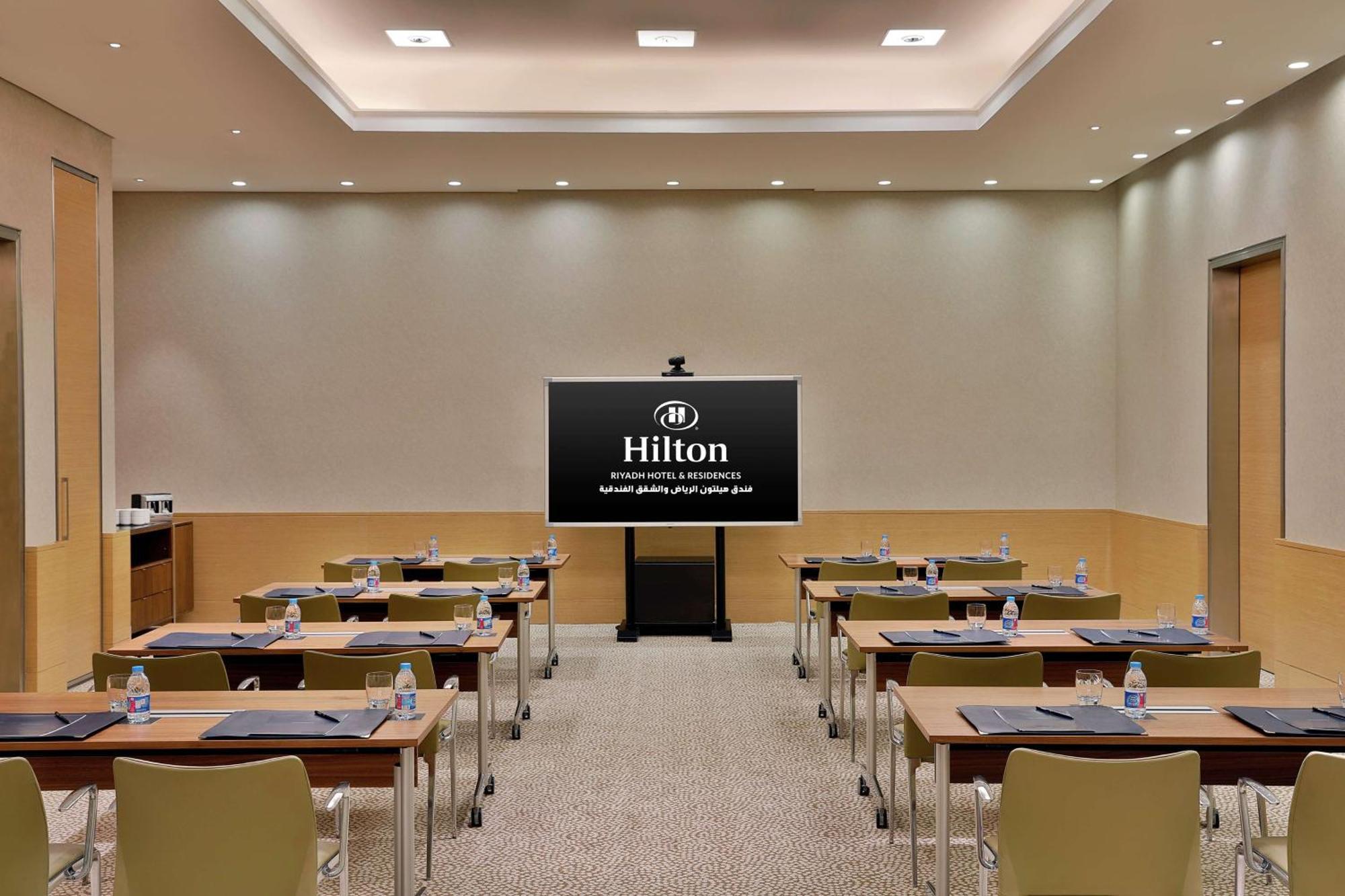 Hilton Riyadh Hotel & Residences Eksteriør billede The photo shows a conference or meeting room set up for a presentation. There are several tables arranged in a classroom style, each equipped with chairs. On each table, there are notepads and water bottles. At the front of the room is a large screen