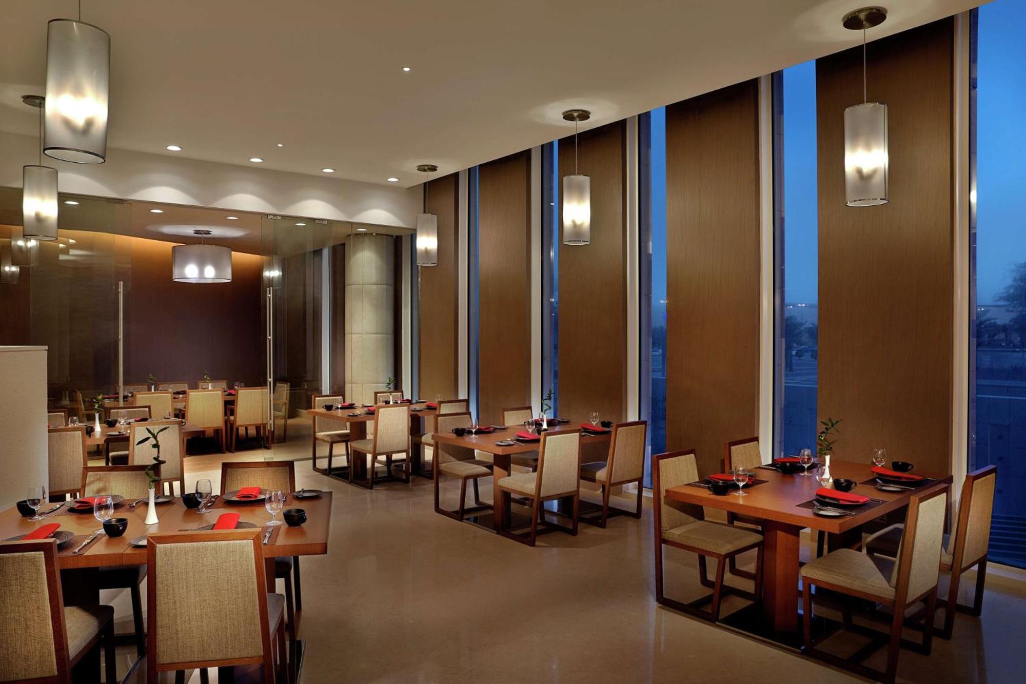 Hilton Riyadh Hotel & Residences Eksteriør billede The image depicts a modern restaurant interior. It features several dining tables arranged neatly, each set with plates and utensils. The room has large windows with blinds partially drawn, allowing some ambient light to filter in, while evening ligh
