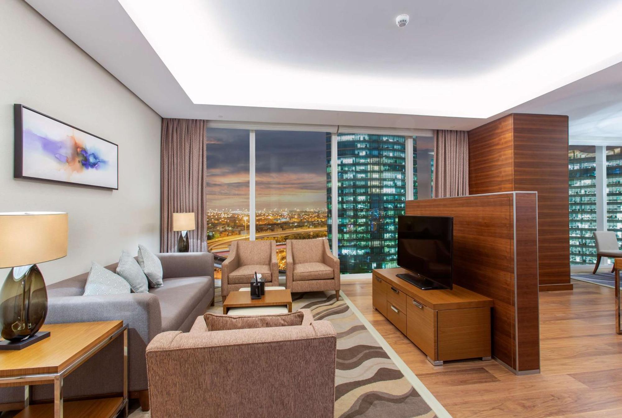 Hilton Riyadh Hotel & Residences Eksteriør billede The photo shows a modern living room in a stylish apartment. The space features a comfortable seating arrangement with a light gray sofa and two brown armchairs, complemented by a wooden coffee table. There is a large flat-screen TV mounted on a wood
