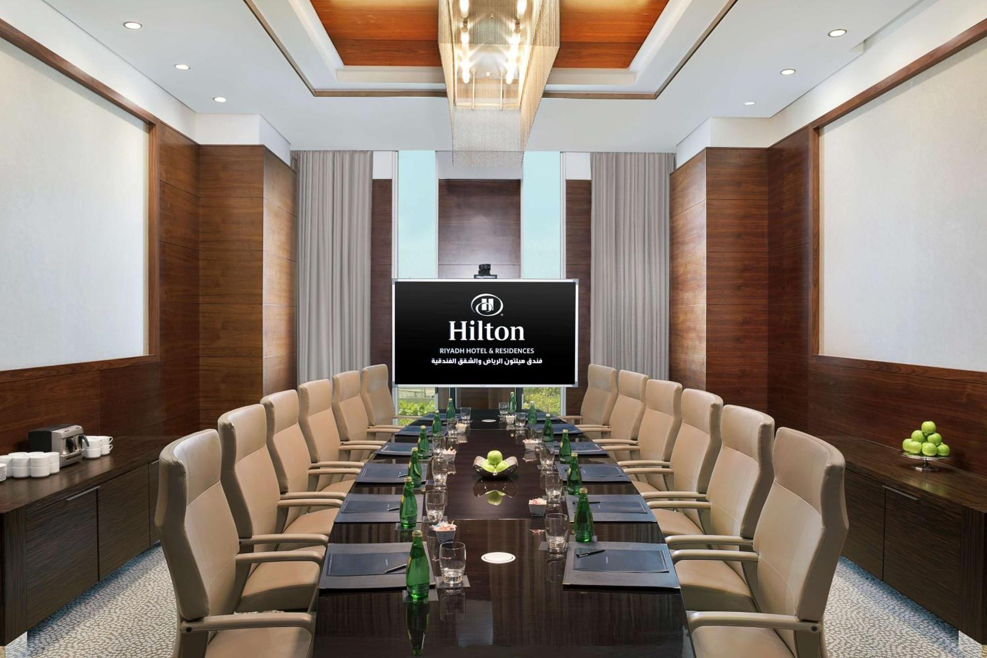 Hilton Riyadh Hotel & Residences Eksteriør billede The photo shows a modern conference room within a Hilton hotel. There is a large wooden table surrounded by several comfortable chairs. At the head of the table is a large screen displaying the Hilton logo. The room is well-lit with stylish overhead 