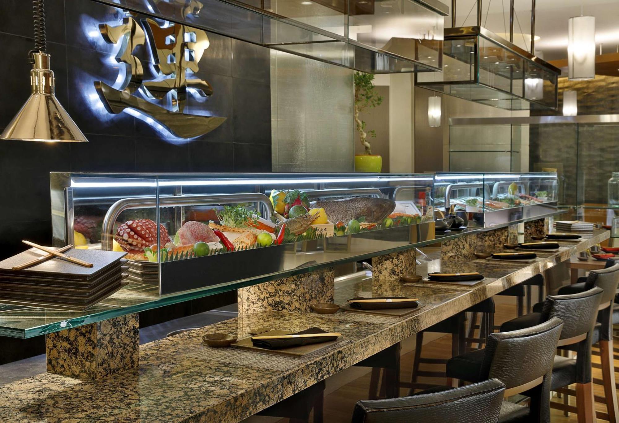 Hilton Riyadh Hotel & Residences Eksteriør billede The photo shows an interior view of a sushi restaurant. There is a long, sleek sushi bar with a glass display case filled with various sushi and sashimi offerings. The counter is made of polished stone, and there are several dark chairs arranged neat